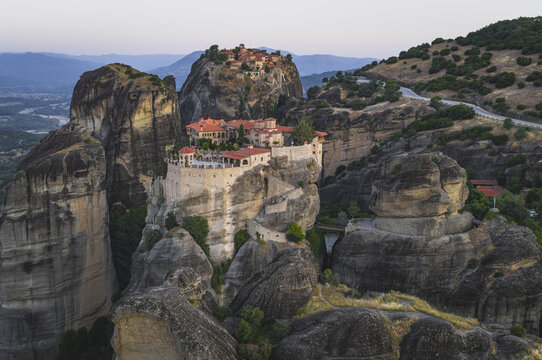 Aerial view of the historic Meteora monasteries perched on massive rock pillars with stone walls and red rooftops under a soft evening sky in Trikala, Greece.
