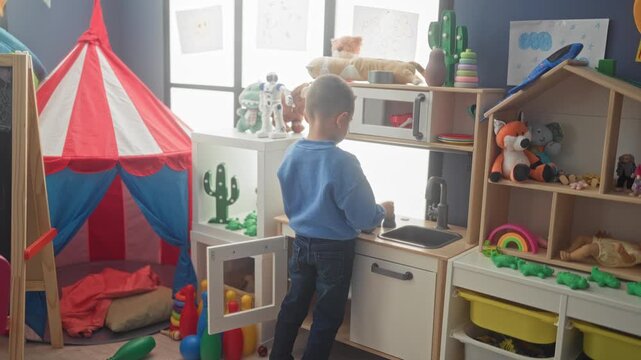 Hispanic boy turns faucet and pretends to wash a toy at a wooden play kitchen in playroom; playful imagination.