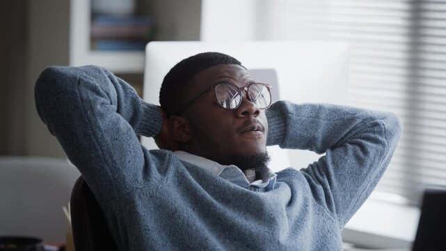 Close up of young adult African American man wearing glasses working on computer, leaning back in office chair with hands behind head to rest. Clip ideal for corporate wellness or IT recruitment ads