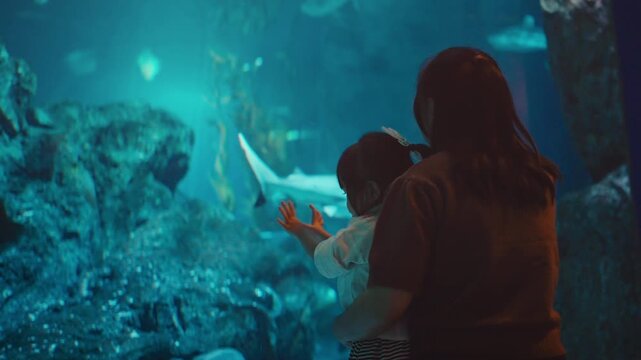Silhouette of mother holding toddler girl looking at sharks and stingrays in large blue aquarium tank. Family travel education and marine life exploration concept. Amazing underwater
