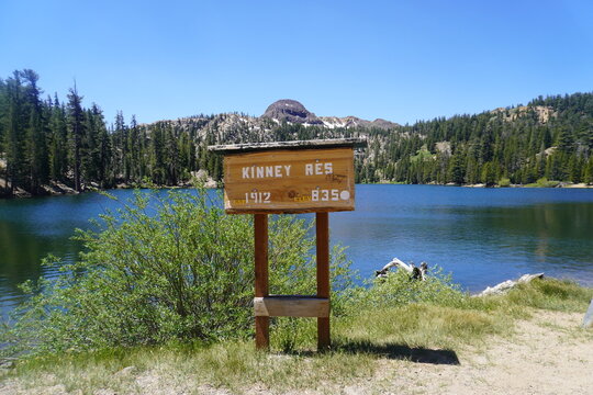 A wide, eye-level shot of a rustic wooden sign marking "KINNEY RES." positioned on a grassy bank overlooking a pristine alpine reservoir. The sign displays carved lettering and elevation details, stan