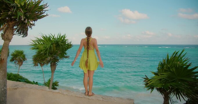 Happy woman in summer dress stands on stone wall overlooking sea in Tulum Mexico