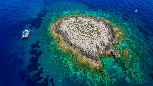Aerial view of a small rocky islet with a stone pillar surrounded by crystal clear turquoise water and a boat nearby off the coast of Vis Split-Dalmatia County, Croatia.