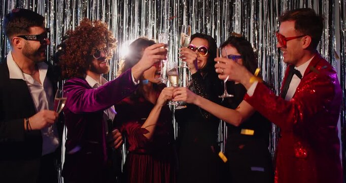 Group of stylish friends in elegant outfits raising champagne glasses for toast at glamorous New Year party. People in festive mood celebrating, laughing and enjoying night with sparkling decorations.