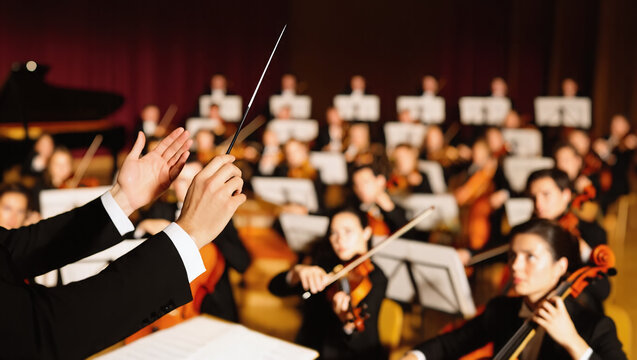 Closeup conductor hands mid-performance, holding a baton high above a professional orchestra background with violinists and cellists in formal attire. Graceful leadership guiding a musical masterpiece