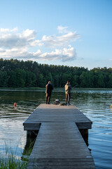 Fototapeta premium Two male friends enjoying a peaceful afternoon fishing from a wooden pier on a calm lake bordered by a dense pine forest under a blue sky.