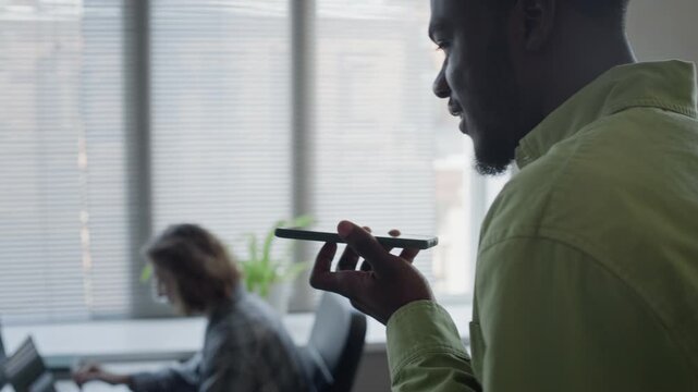 Tracking shot of young adult African American man recording voice message on smartphone in office with colleague working in background. For mobile app development and business communication ads
