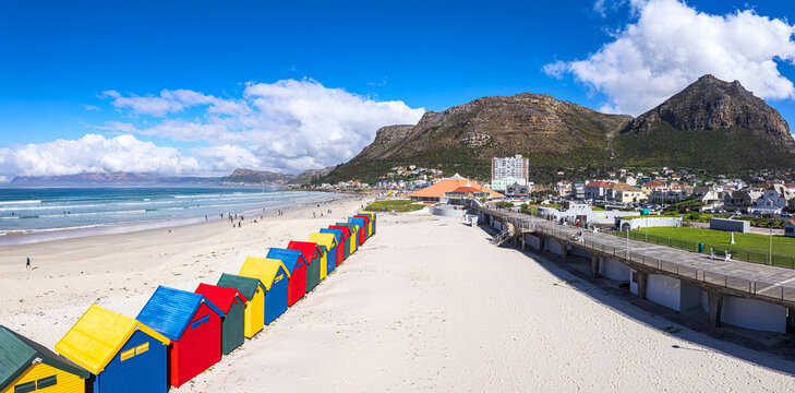 Aerial view of the iconic colorful beach huts on the sandy shore of Muizenberg Beach with mountains and a promenade in Cape Town, Western Cape, South Africa.