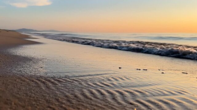 Low angle close-up of small ocean waves washing onto a sandy beach at sunset. Golden hour light reflects on water and wet sand with a clear horizon and distant hills in the background.