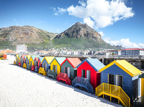 Aerial view of the row of colorful beach huts at Muizenberg Beach on white sand with rugged mountains under a blue sky in Cape Town, Western Cape, South Africa.