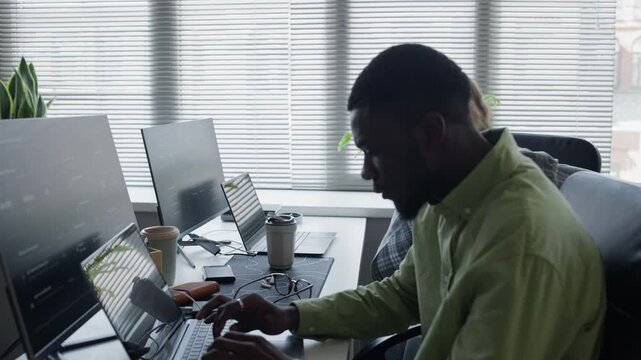 Tracking shot of young Caucasian woman walking to desk and sitting next to young African American man to write code on laptops in office. For tech startup commercials and software development videos