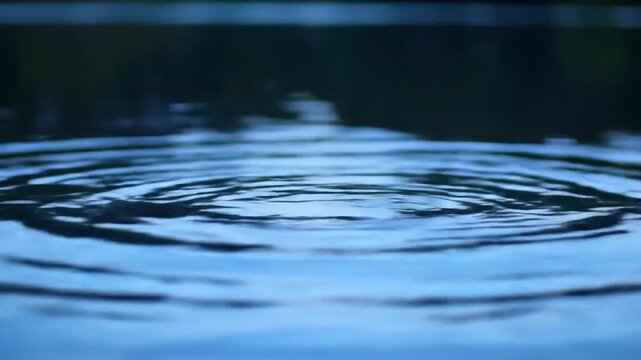 Close-up of circular ripples expanding across a calm dark blue water surface, showcasing concentric wave patterns and liquid texture in low light cinematic environment.