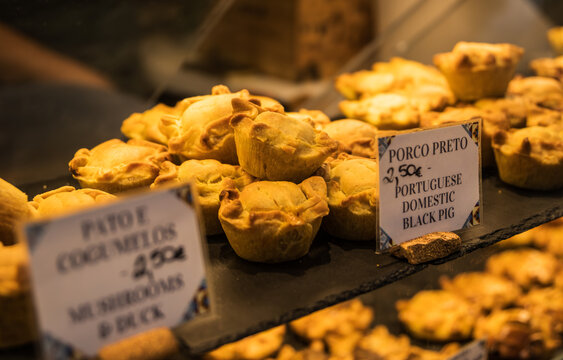 Primer plano de pasteles de masa salada rellenos de cerdo negro, pato y setas en un mostrador con carteles de precios. Aperitivos tradicionales listos para comer en un mercado de Portugal.