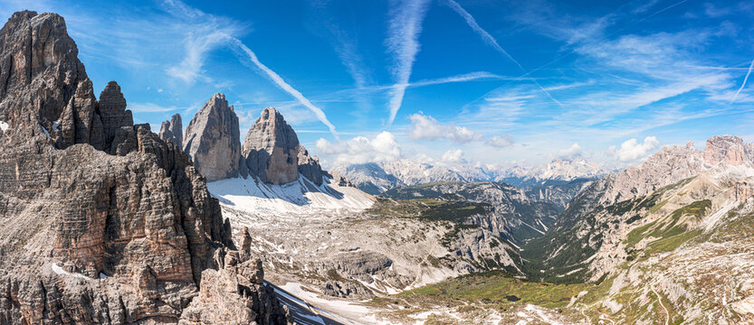 Aerial view of the Tre Cime di Lavaredo peaks with jagged rock formations and a deep valley under a blue sky with contrails in Sexten, Trentino-Alto Adige S, Italy.