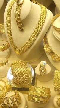 Close-up of ornate gold jewelry displayed in a shop window, showing necklaces, bracelets, rings and bangles with a slight camera shift across the luxury showcase