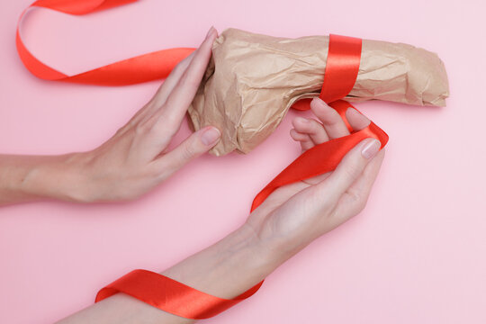 Female hands wrapping gift silicon sextoy vibrator with red ribbon on pink background