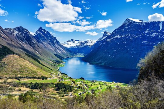 The scene shows village of Urke in the Hjorundfjord in Norway surrounded by tall mountains and green fields. 