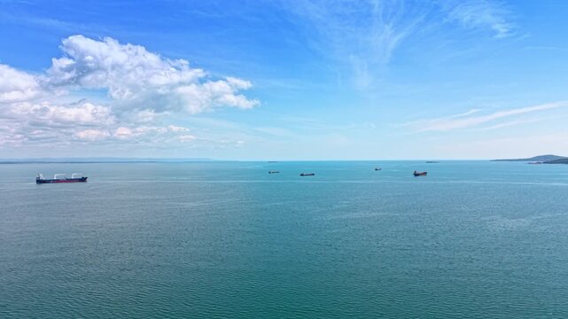 Fleet of cargo ships scattered across Burgas Bay on a clear day. Aerial panorama of multiple vessels on teal water with island and cumulus clouds.