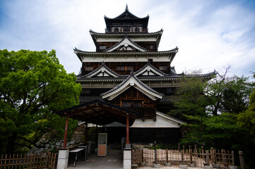 Fototapeta premium The historic main wooden keep of Hiroshima Castle surrounded by green trees under a cloudy sky, Japan