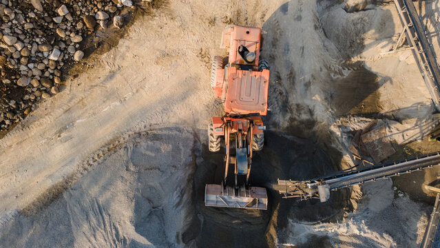 Aerial view of an orange wheel loader moving sand in a quarry with conveyor belts and rock piles in Karanglewas, Central Java, Indonesia.