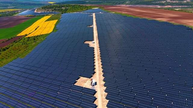 Massive solar farm divided by central access road. Symmetrical panel rows flanking dirt path with rapeseed patches, cropland and distant hills.