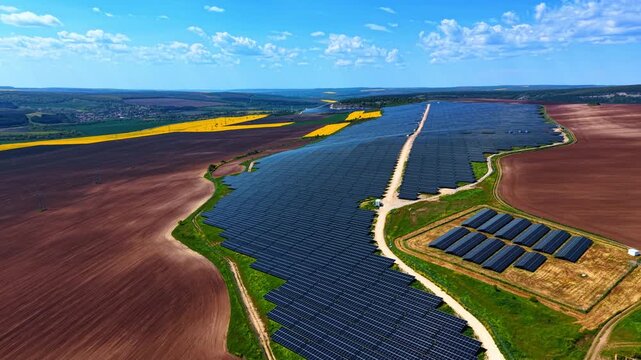 Solar panel arrays stretching between plowed farmland and rapeseed crops. Elongated rows curving along terrain with yellow fields, brown earth and village on horizon.