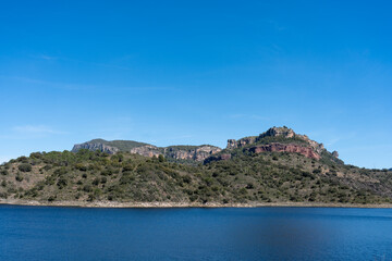 Fototapeta premium Wide, expansive shot of a tranquil natural reserve where a pristine dark water body meets rolling green shrubland, backed by towering, flat-topped rocky mountains on a sunny day.