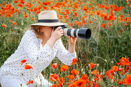 Crouching woman photographer in straw hat shooting with telephoto lens inside poppy field. Artistic process, outdoor work, nature photography masterclass and creative lifestyle hobby.
