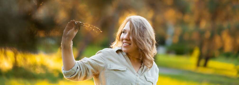 Contemplative woman inspects sunglasses in sunlit park. Sophisticated summer style, dreamy afternoon mood.