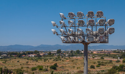 Large stadium floodlight tower with multiple lamp heads rising above a rural Spanish landscape with mountains and town in background, symbolising energy consumption and light pollution © IgorsPerspective