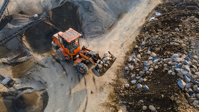 Aerial view of an orange wheel loader carrying large rocks at a quarry site with sand piles and conveyor belts in Karanglewas, Central Java, Indonesia.