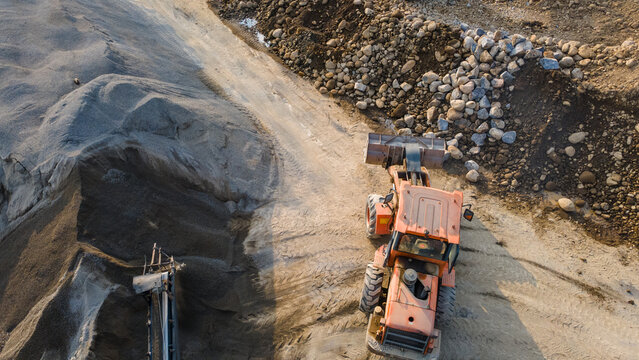 Aerial view of an orange wheel loader moving earth and rocks near large piles of sand and gravel at a construction site in Karanglewas, Central Java, Indonesia.