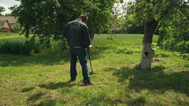 Treasure hunter using metal detector in grassy field near trees, searching for hidden objects in a rural outdoor setting