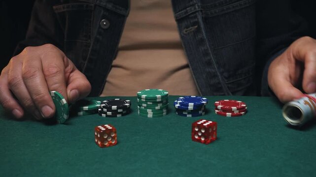 Male gambler places green poker chip on table surrounded by colorful chips and dice in a dimly lit casino setting