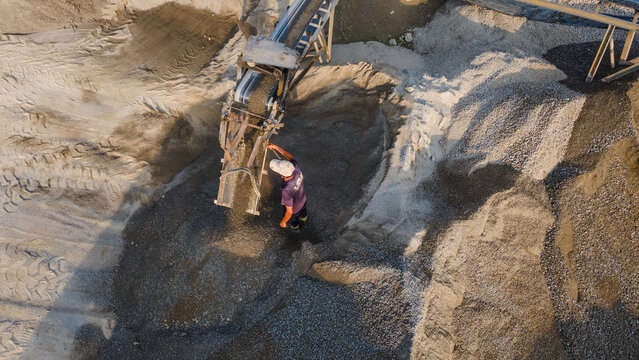 Aerial view of a worker standing by a conveyor belt pouring gravel onto a large pile at a sand quarry with visible tire tracks Karanglewas, Central Java, Indonesia.