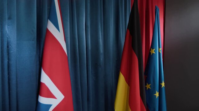 International flags representing United Kingdom, Germany, and European Union hanging against dark curtain during political summit. Clip is suitable for news broadcasts and political commentary