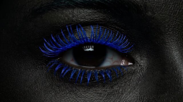 Close-up of a dark-skinned face as one eye opens with vivid blue eyelashes and bold eyeliner in low light