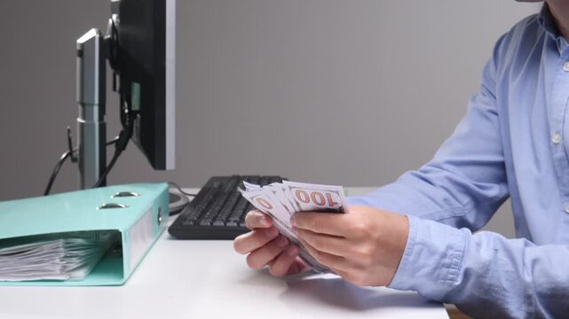 Hands of a person wearing a blue shirt sitting at an office desk, accurately counting a stack of us hundred dollar bills, managing personal finances or business earnings