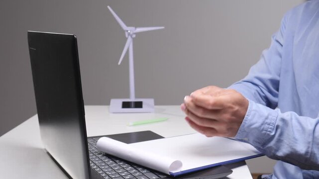 Person's hands crumpling a piece of paper over an open notebook and laptop on a desk, with a miniature wind turbine model visible in the background, symbolizing challenges in renewable energy work