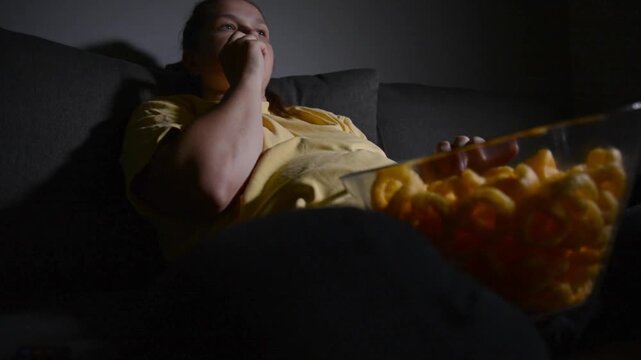 Young woman sitting on a sofa and eating corn rings while watching a movie. Cozy home entertainment scene focused on snacking, relaxation and leisure.