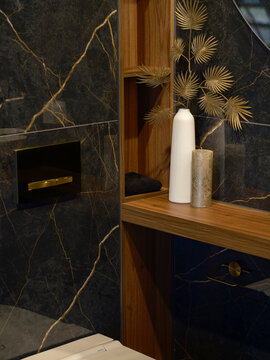 Detail of a dark veined marble bathroom with a gold flush plate, a wood shelf niche with a vase and candle and a blue stone cabinet.