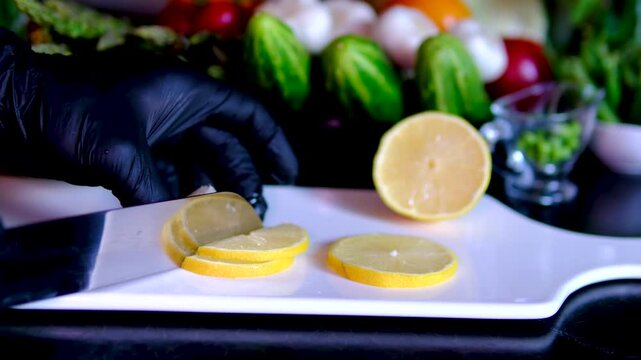 Chef in black gloves slicing fresh lemon into thin rounds on white porcelain board close up