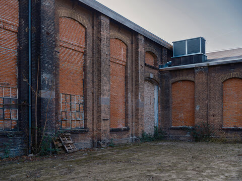 Exterior of an abandoned brick factory with tall arched windows and a rooftop air handling unit beside an empty courtyard.