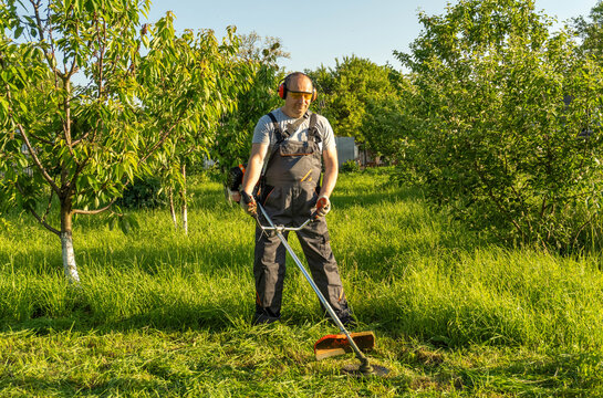 A worker stands in a garden, trimming grass with a grass trimmer on a sunny day. He wears safety gear and works among trees and high grass. The scene shows a typical outdoor task