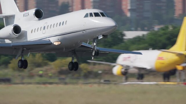 Close up tracking shot of a white private business jet approaching and touching down on a runway, landing gear extended, urban buildings visible in background, daylight.