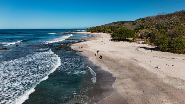 Aerial view of the tropical coastline with people horseback riding on the sandy beach and waves crashing against the shore in Santa Teresa, Puntarenas Province, Costa Rica.