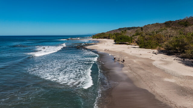 Aerial view of people riding horses along the sandy shoreline of a tropical beach with crashing waves and lush forest in Santa Teresa, Puntarenas Province, Costa Rica.
