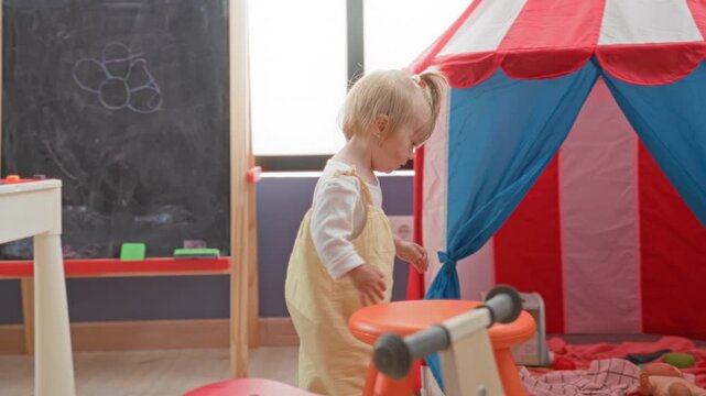 Toddler girl holding a doll and pulling the play tent curtain while standing near a toy bike and stool in a building; curiosity innocence.