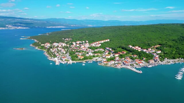 Aerial view of Klimno, Krk Island, Croatia, showcasing its charming marina, red-roofed houses, and lush green hills under a clear blue sky.