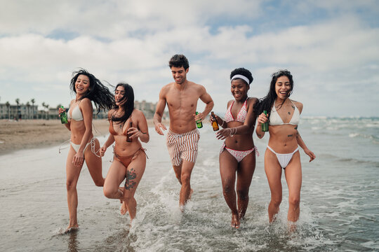 Diverse group of young friends running laughing splashing water on beach enjoying summer party vacation